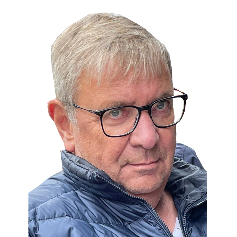 Ein Mann mit grauem Haar, Brille und blauer Jacke schaut in die Kamera